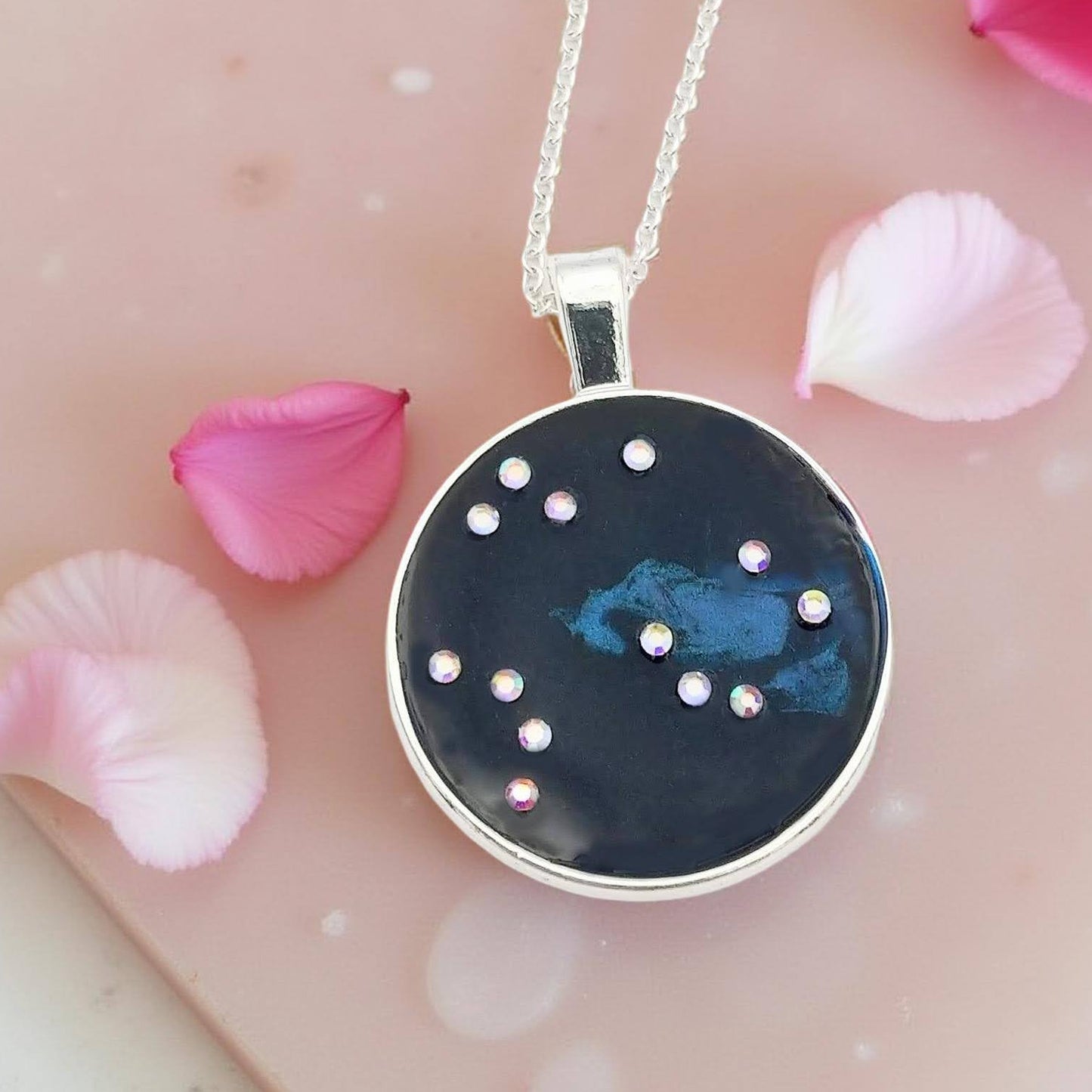 Dark blue with iridescent sparkling swirls of pinks, purples, silver, and monochromatic shades of blue enamel. With handset Aurora Borealis rhinestones to represent the star of the constellation of Aquarius. The pendant is 1 inch silver toned with a 1/4th inch bail on a silver toned chain.