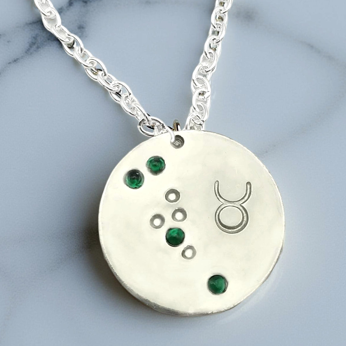 May Taurus necklace pendant with hand set emerald colored glass rhinestones forming the constellation of Taurus. Hand stamped with the zodiac symbol for Taurus, on a 3/4th of an inch silver toned circle hanging from a silver toned chain.