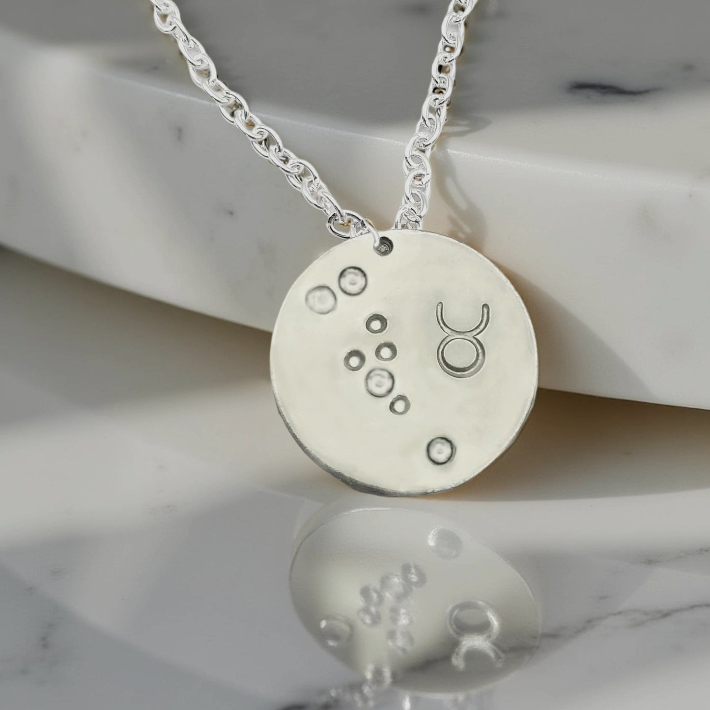 April Taurus necklace pendant with hand set diamond colored glass rhinestones forming the constellation of Taurus. Hand stamped with the zodiac symbol for Taurus, on a 3/4th of an inch silver toned circle hanging from a silver toned chain.