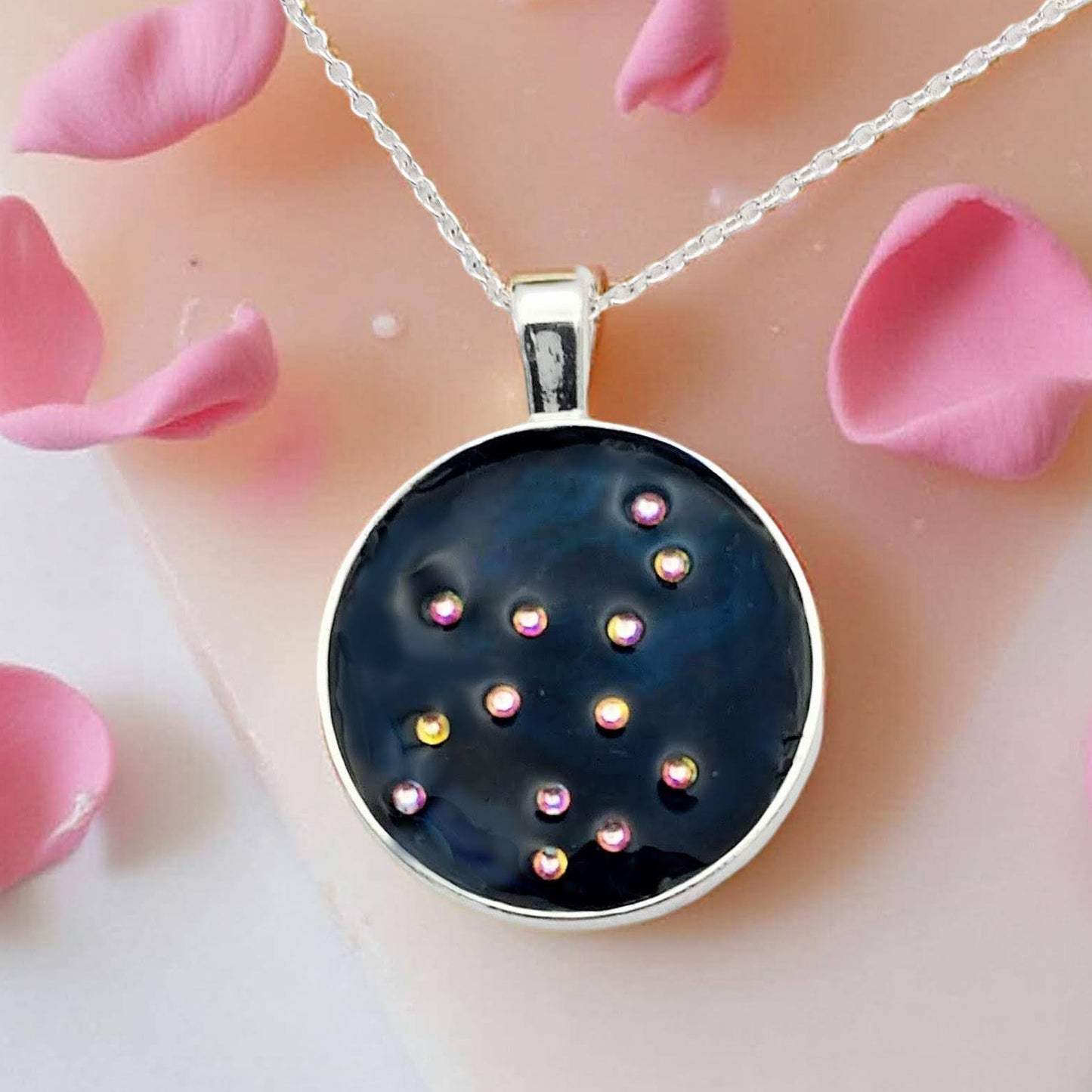 Dark blue with iridescent sparkling swirls of pinks, purples, silver, and monochromatic shades of blue enamel. With handset Aurora Borealis rhinestones to represent the star of the constellation of Virgo. The pendant is 1 inch silver toned with a 1/4th inch bail on a silver toned chain.