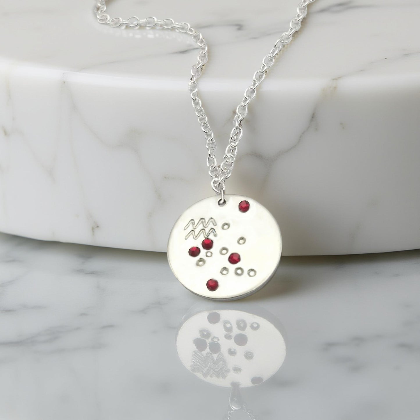 January Aquarius necklace pendant with hand set garnet colored glass rhinestones forming the constellation of Aquarius. Hand stamped with the zodiac symbol for Aquarius, on a 3/4th of an inch silver toned circle hanging from a silver toned chain.