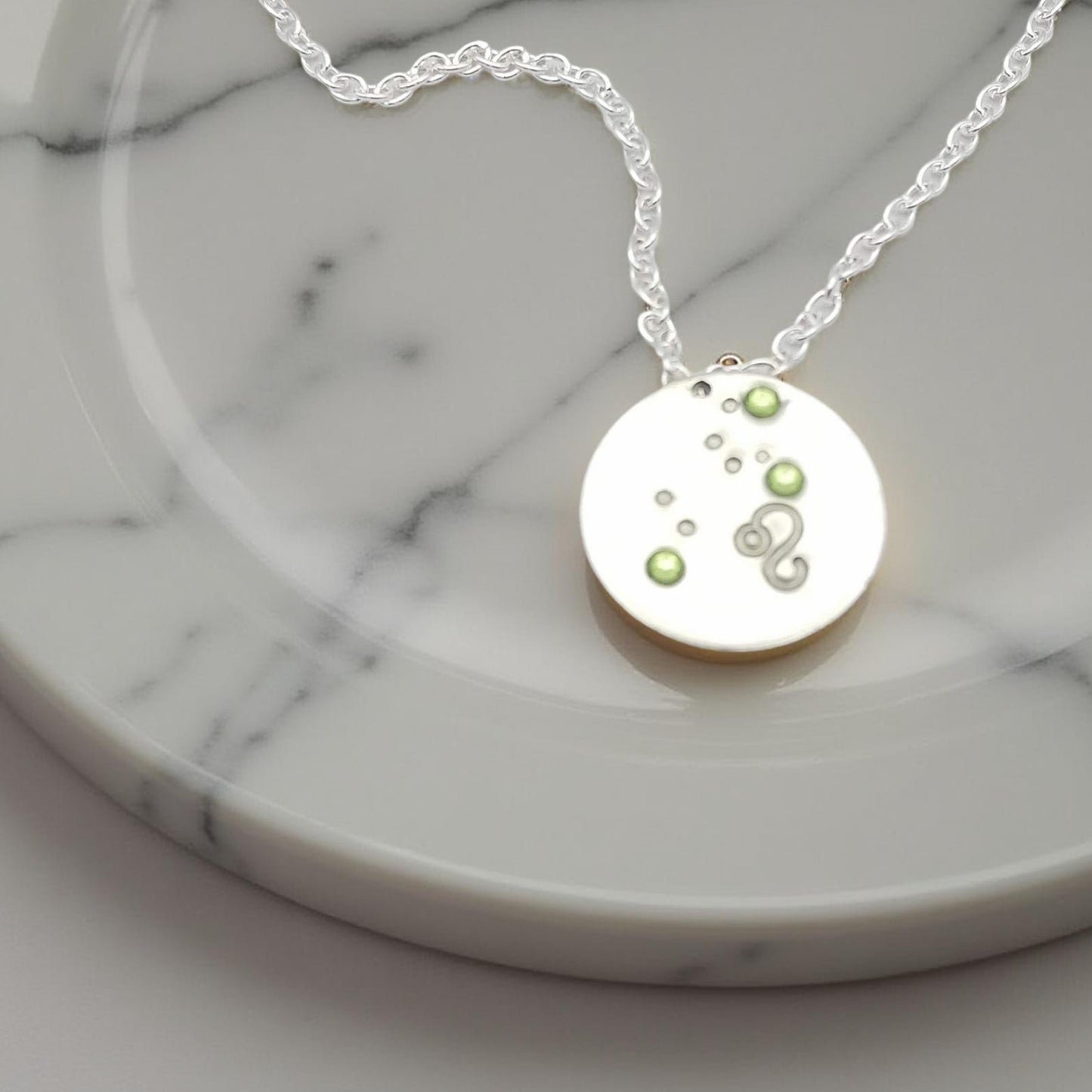 August Leo necklace pendant with hand set peridot colored glass rhinestones forming the constellation of Leo. Hand stamped with the zodiac symbol for Leo, on a 3/4th of an inch silver toned circle hanging from a silver toned chain.