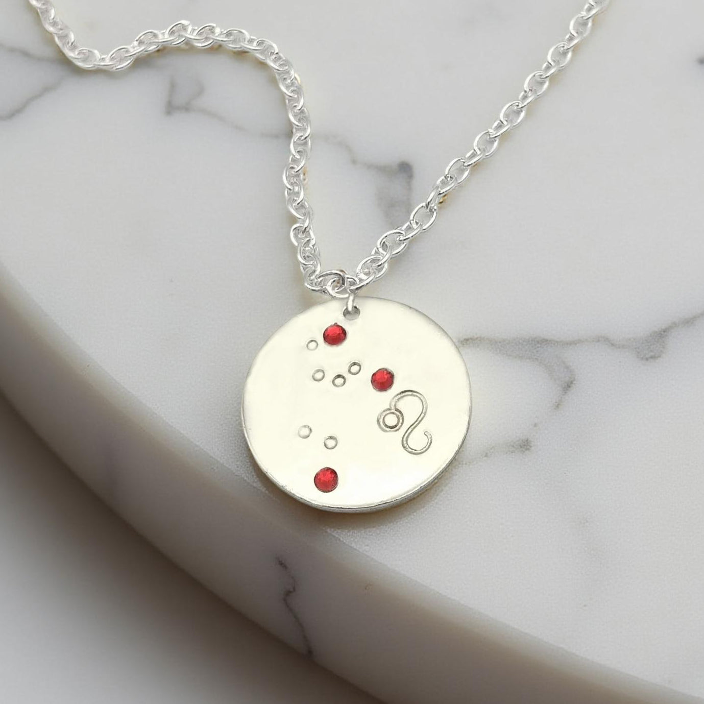 July Leo necklace pendant with hand set ruby colored glass rhinestones forming the constellation of Leo. Hand stamped with the zodiac symbol for Leo, on a 3/4th of an inch silver toned circle hanging from a silver toned chain.