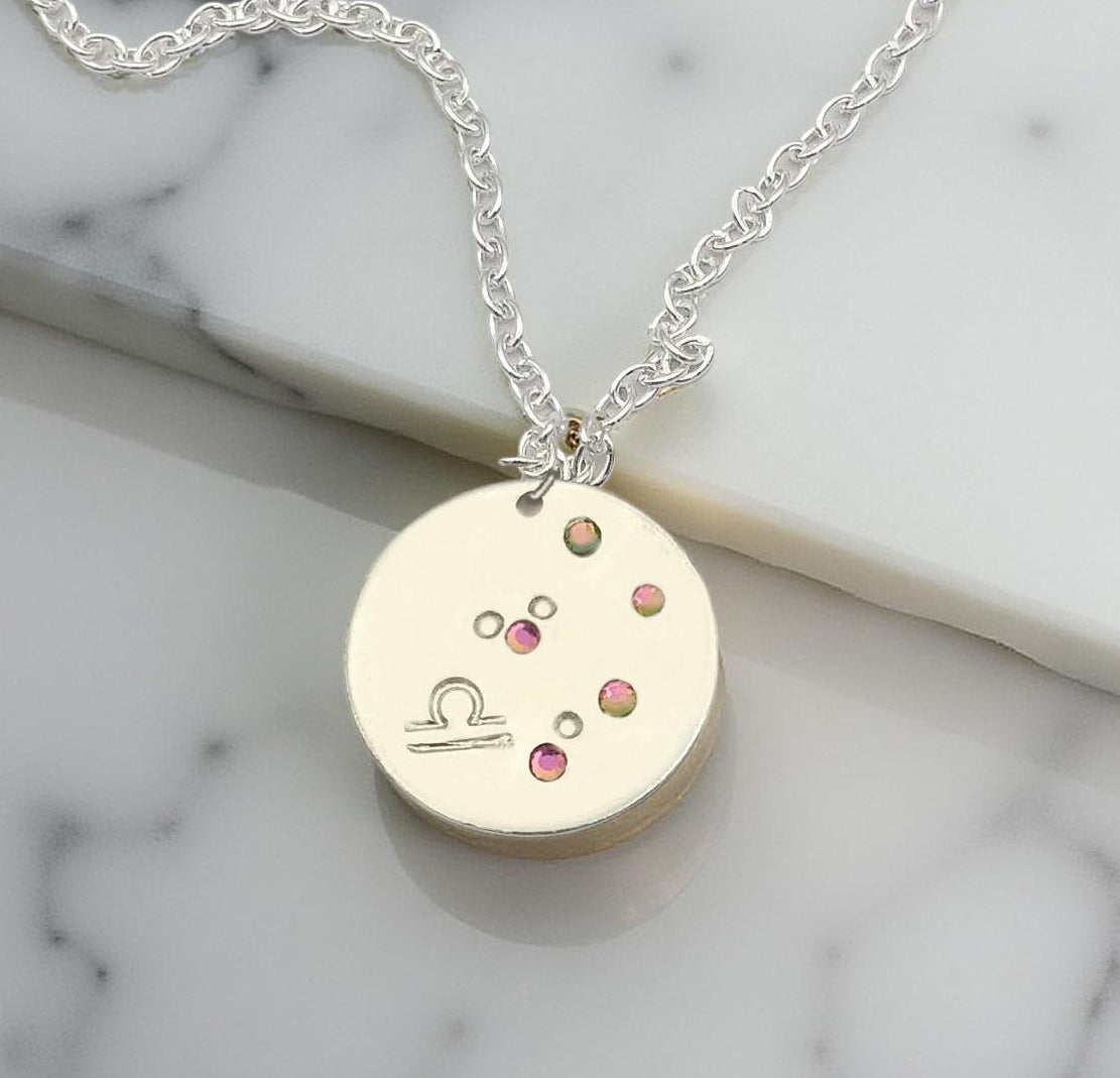 October Libra necklace pendant with hand set tourmaline colored glass rhinestones forming the constellation of Libra. Hand stamped with the zodiac symbol for Libra, on a 3/4th of an inch silver toned circle hanging from a silver toned chain.