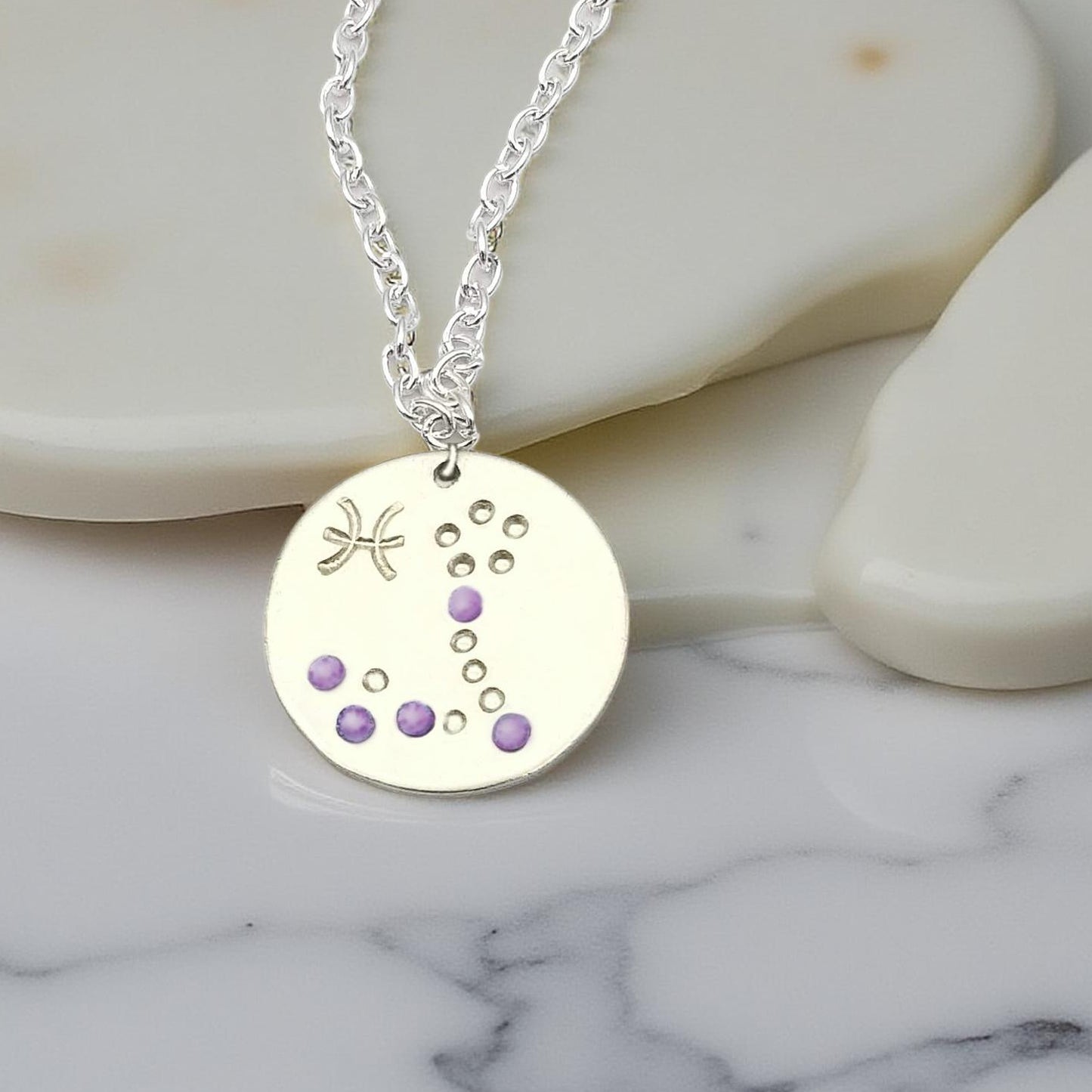February Pisces necklace pendant with hand set amethyst colored glass rhinestones forming the constellation of Pisces. Hand stamped with the zodiac symbol for Pisces, on a 3/4th of an inch silver toned circle hanging from a silver toned chain.