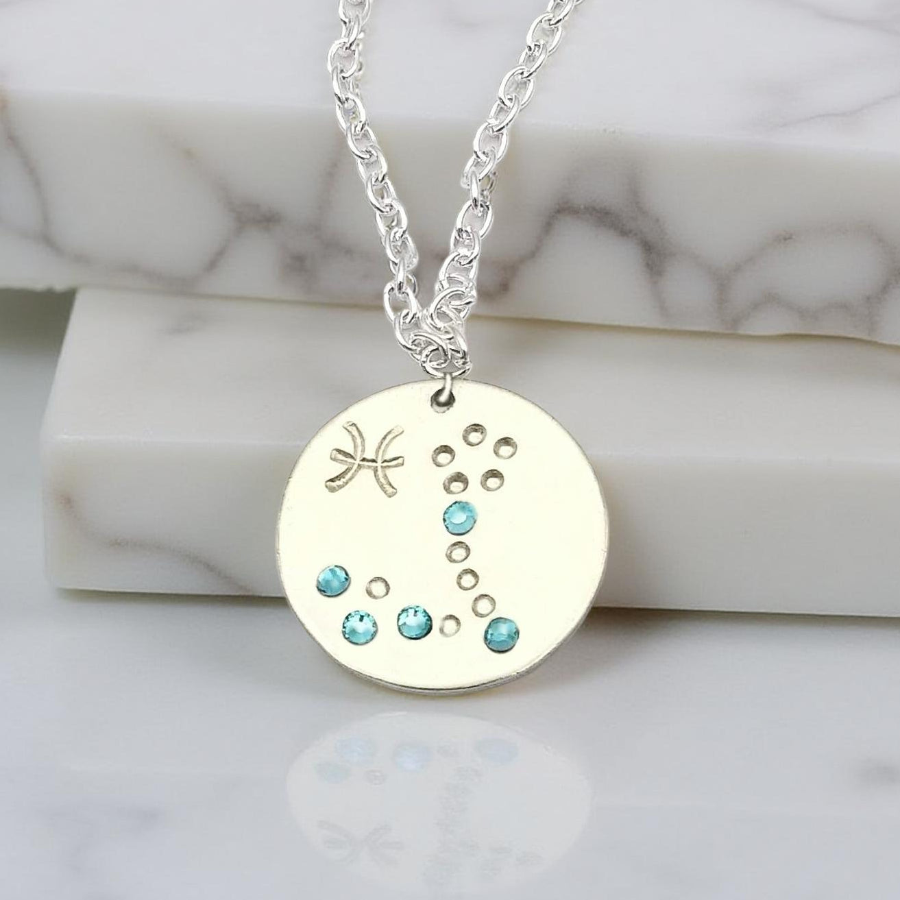 March Pisces necklace pendant with hand set aquamarine colored glass rhinestones forming the constellation of Pisces. Hand stamped with the zodiac symbol for Pisces, on a 3/4th of an inch silver toned circle hanging from a silver toned chain.