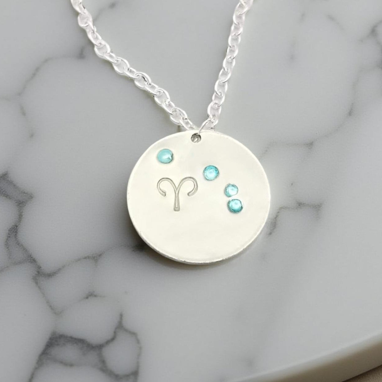 March Aries necklace pendant with hand set aquamarine colored glass rhinestones forming the constellation of Aries. Hand stamped with the zodiac symbol for aries, on a 3/4th of an inch silver toned circle hanging from a silver toned chain.