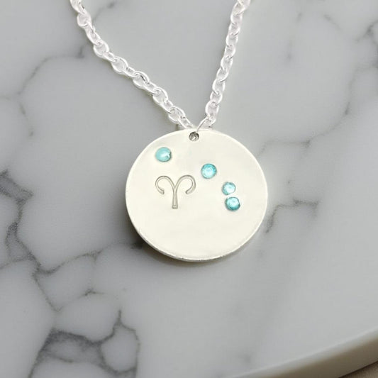 March Aries necklace pendant with hand set aquamarine colored glass rhinestones forming the constellation of Aries. Hand stamped with the zodiac symbol for aries, on a 3/4th of an inch silver toned circle hanging from a silver toned chain.