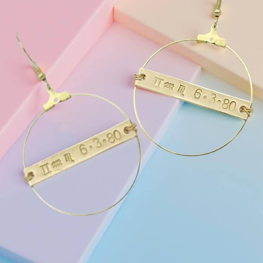 Gold toned 1 1/2 inch diameter thin hoop earrings on fish hooks.  With a hand stamped 1 1/2 inch  X 1/4th inch  brass/gold toned plaque with zodiac symbols of customizable rising, sun and moon signs along with your date of birth.  Centered in the middle of the hoop with wire wrapping.