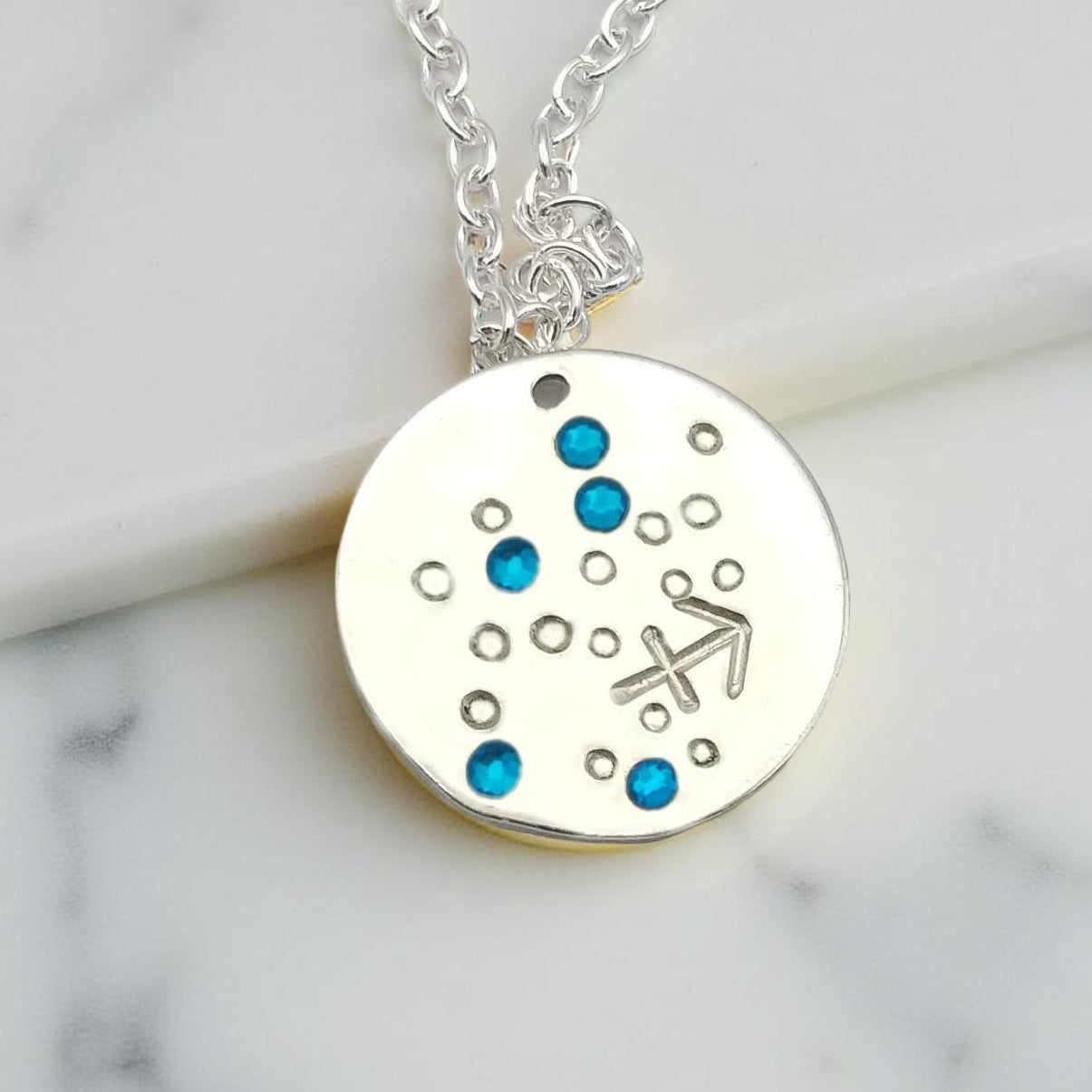 December Sagittarius necklace pendant with hand set tanzanite colored glass rhinestones forming the constellation of Sagittarius. Hand stamped with the zodiac symbol for Sagittarius, on a 3/4th of an inch silver toned circle hanging from a silver toned chain.