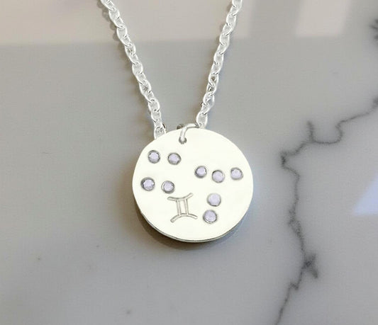 June Gemini necklace pendant with hand set alexandrite colored glass rhinestones forming the constellation of Gemini. Hand stamped with the zodiac symbol for Gemini, on a 3/4th of an inch silver toned circle hanging from a silver toned chain.