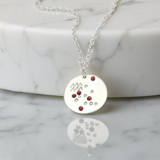January Aquarius necklace pendant with hand set garnet colored glass rhinestones forming the constellation of Aquarius.  Hand stamped with the zodiac symbol for Aquarius, on a 3/4th of an inch silver toned circle hanging from a silver toned chain.
