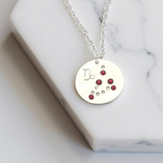 January Capricorn necklace pendant with hand set garnet colored glass rhinestones forming the constellation of Capricorn. Hand stamped with the zodiac symbol for Capricorn, on a 3/4th of an inch silver toned circle hanging from a silver toned chain.