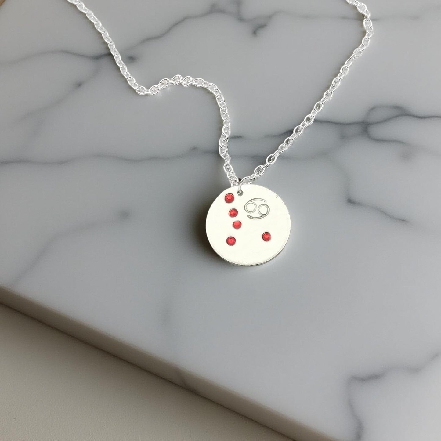 July Cancer necklace pendant with hand set ruby colored glass rhinestones forming the constellation of Cancer. Hand stamped with the zodiac symbol for Cancer, on a 3/4th of an inch silver toned circle hanging from a silver toned chain.