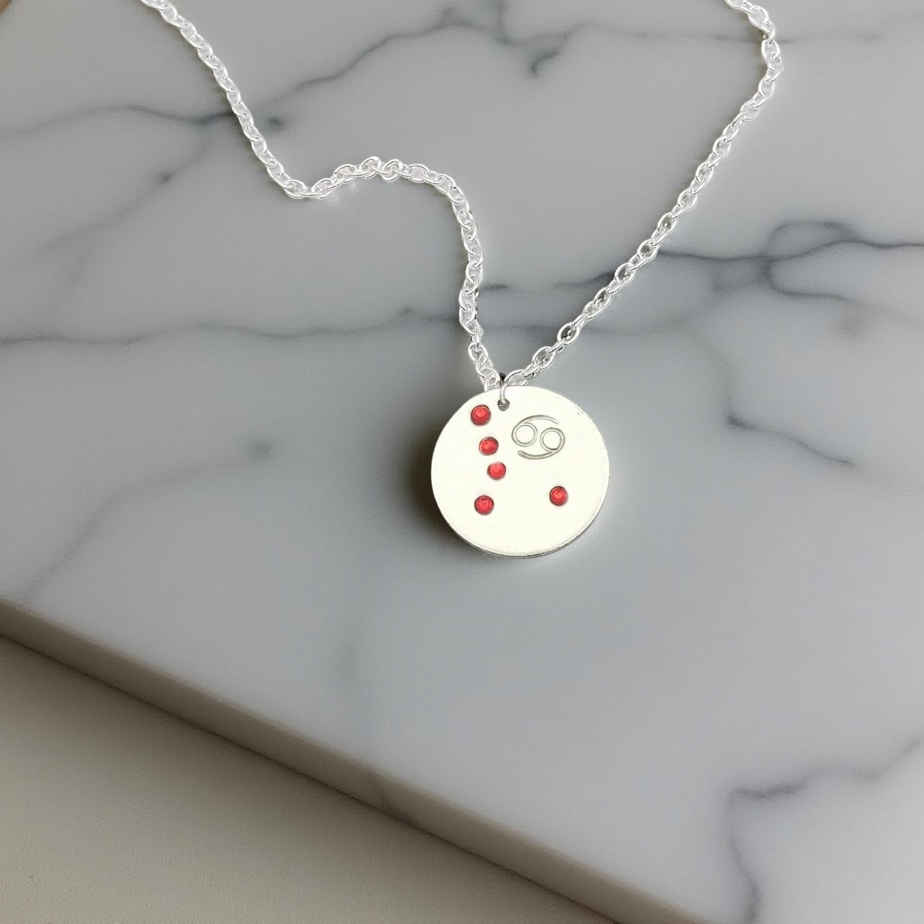 July Cancer necklace pendant with hand set ruby colored glass rhinestones forming the constellation of Cancer. Hand stamped with the zodiac symbol for Cancer, on a 3/4th of an inch silver toned circle hanging from a silver toned chain.