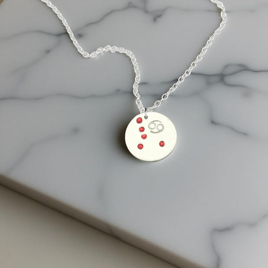 July Cancer necklace pendant with hand set ruby colored glass rhinestones forming the constellation of Cancer. Hand stamped with the zodiac symbol for Cancer, on a 3/4th of an inch silver toned circle hanging from a silver toned chain.