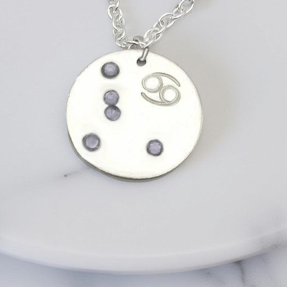 June Cancer necklace pendant with hand set alexandrite colored glass rhinestones forming the constellation of Cancer. Hand stamped with the zodiac symbol for Cancer, on a 3/4th of an inch silver toned circle hanging from a silver toned chain.