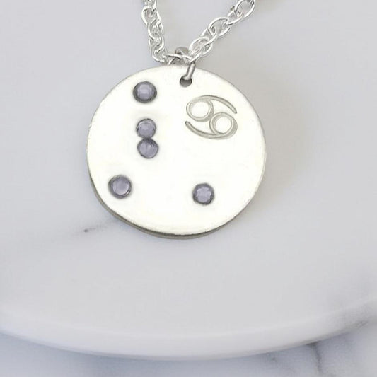 June Cancer necklace pendant with hand set alexandrite colored glass rhinestones forming the constellation of Cancer. Hand stamped with the zodiac symbol for Cancer, on a 3/4th of an inch silver toned circle hanging from a silver toned chain.