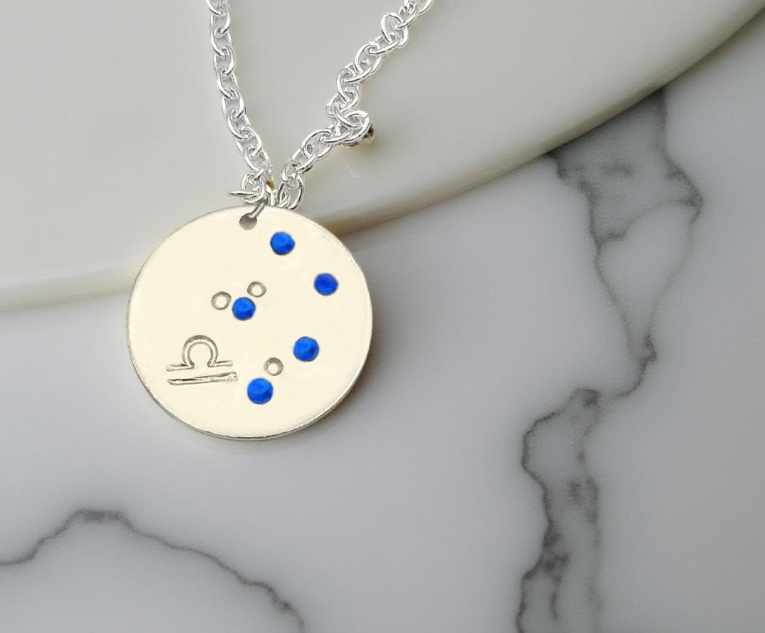 September Libra necklace pendant with hand set sapphire colored glass rhinestones forming the constellation of Libra. Hand stamped with the zodiac symbol for Libra, on a 3/4th of an inch silver toned circle hanging from a silver toned chain.