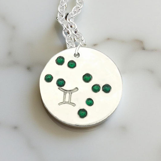 May Gemini necklace pendant with hand set emerald colored glass rhinestones forming the constellation of Gemini. Hand stamped with the zodiac symbol for Gemini, on a 3/4th of an inch silver toned circle hanging from a silver toned chain.