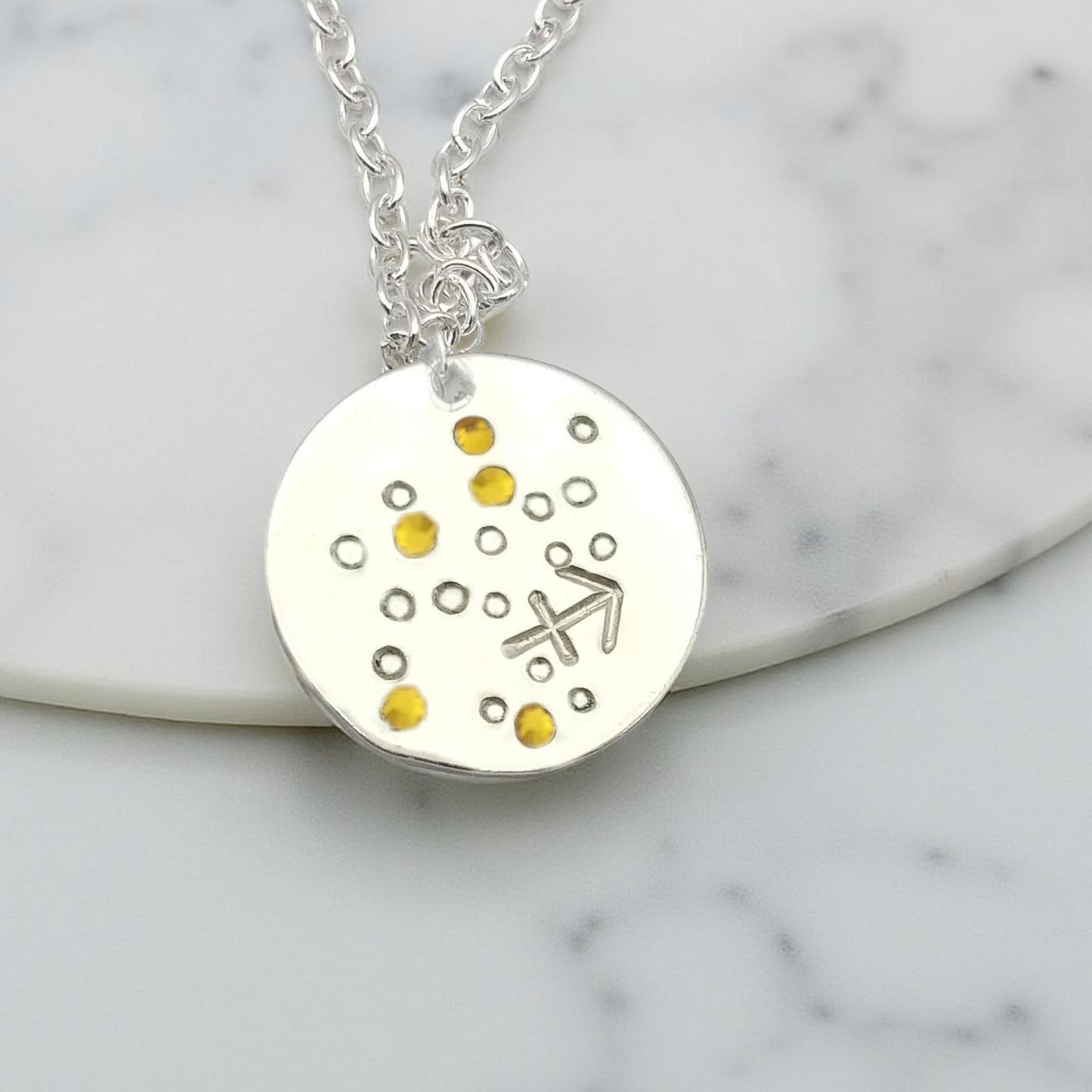 November Sagittarius necklace pendant with hand set citrine colored glass rhinestones forming the constellation of Sagittarius. Hand stamped with the zodiac symbol for Sagittarius, on a 3/4th of an inch silver toned circle hanging from a silver toned chain.