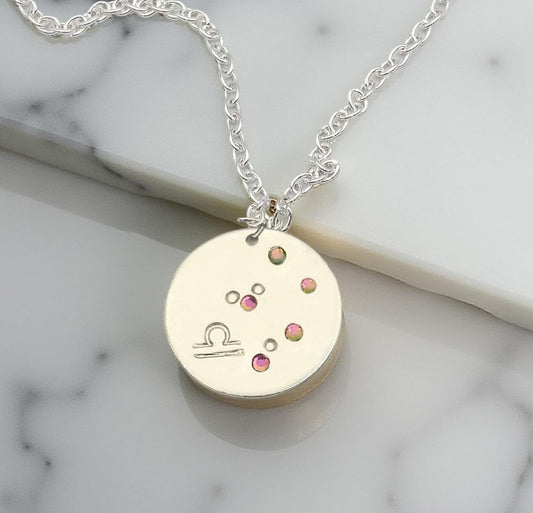 October Libra necklace pendant with hand set tourmaline colored glass rhinestones forming the constellation of Libra. Hand stamped with the zodiac symbol for Libra, on a 3/4th of an inch silver toned circle hanging from a silver toned chain.