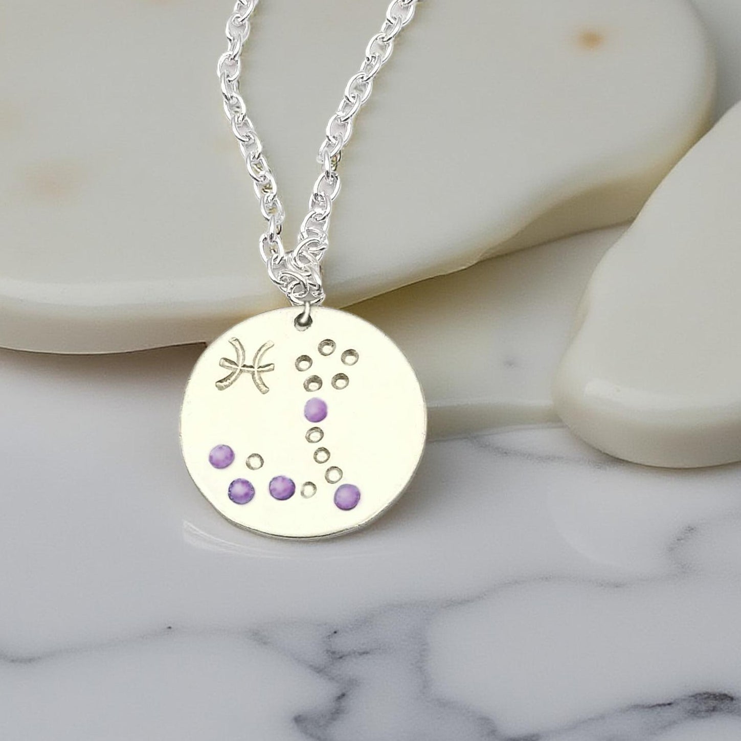 February Pisces necklace pendant with hand set amethyst colored glass rhinestones forming the constellation of Pisces. Hand stamped with the zodiac symbol for Pisces, on a 3/4th of an inch silver toned circle hanging from a silver toned chain.