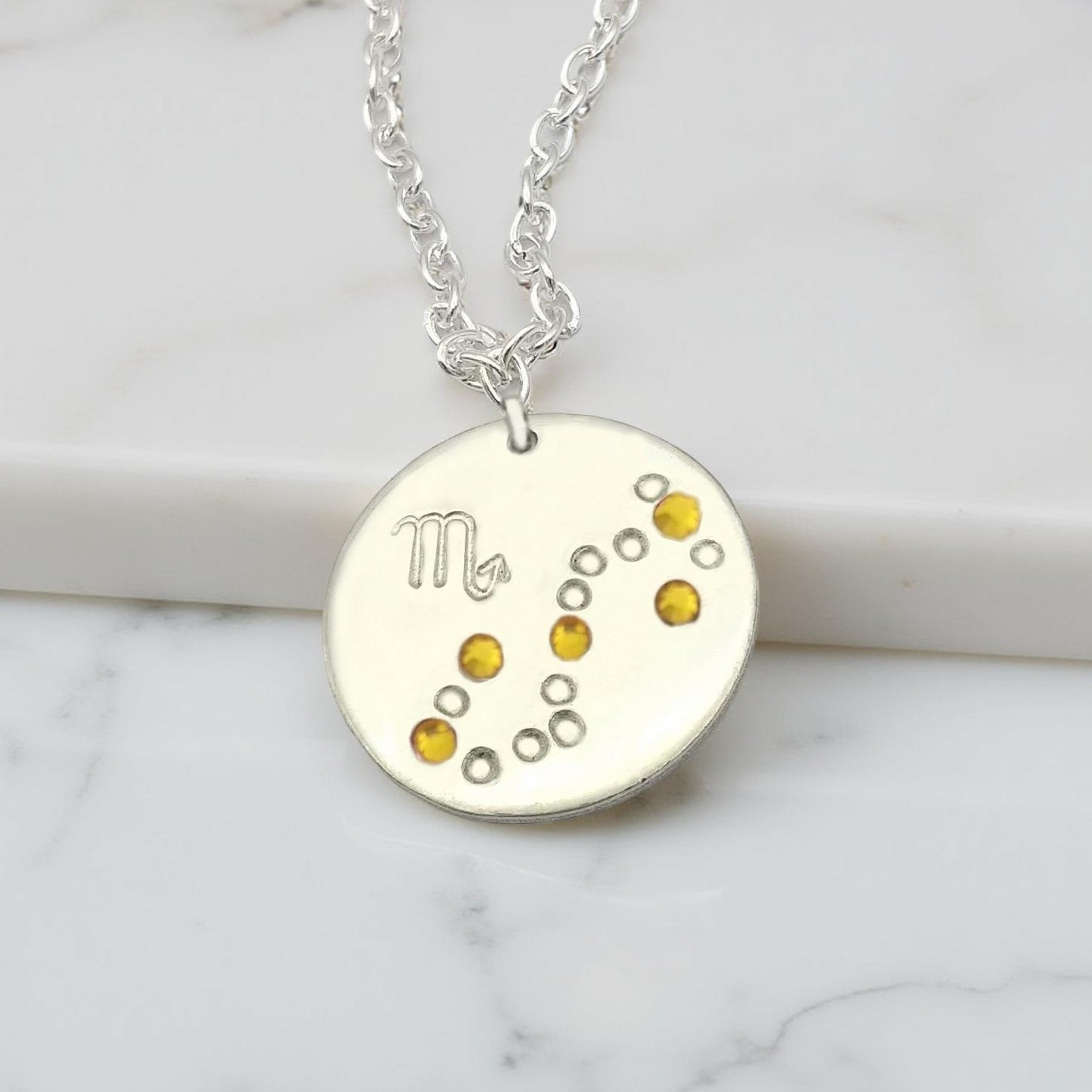 November Scorpio necklace pendant with hand set citrine colored glass rhinestones forming the constellation of Scorpio. Hand stamped with the zodiac symbol for Scorpio, on a 3/4th of an inch silver toned circle hanging from a silver toned chain.